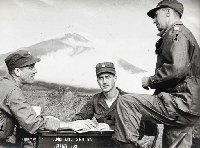 black white photo of 3 men standing by a box with a mountain in the background