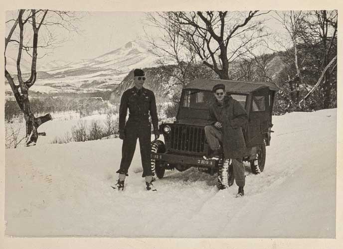 black white photo of 2 men standing in front a jeep in the snow