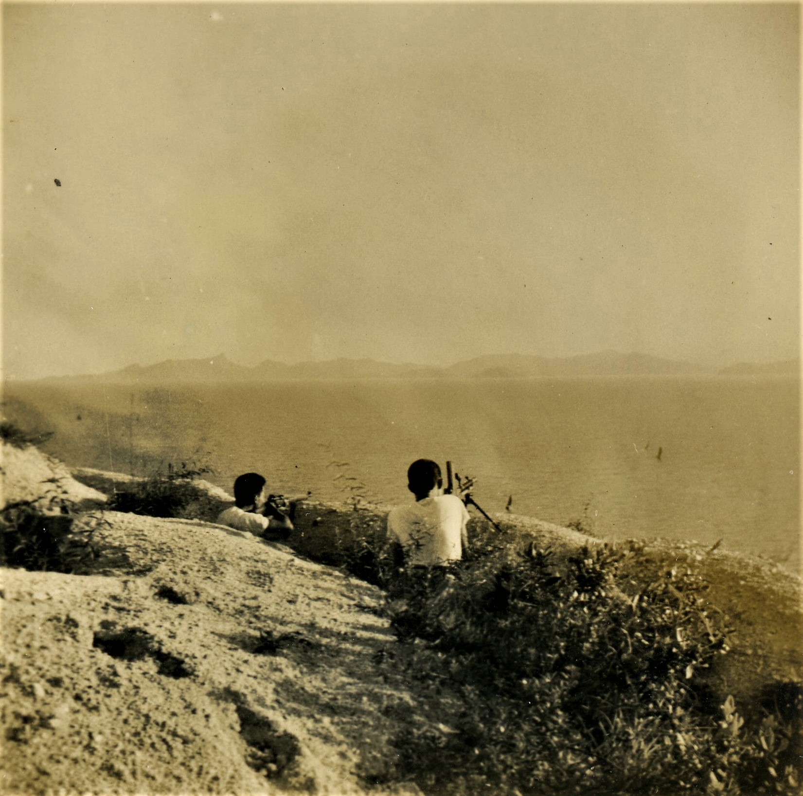 Guerrilla fighters crouching behind a hill overlooking the ocean and ships.