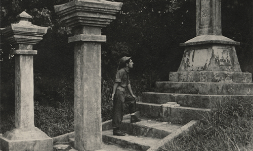 A black-and-white photograph showing a young US Navy Seabee standing on stone steps, looking upward at a Japanese stone memorial monument.