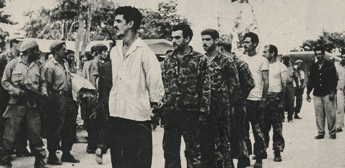 Detail of black and white photo showing prisoners bound and lined up as armed gaurds stand around them