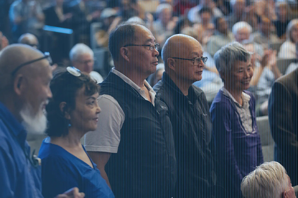 A row of diverse older adults standing attentively in a crowded auditorium, viewed in profile against a blurred background of audience members.