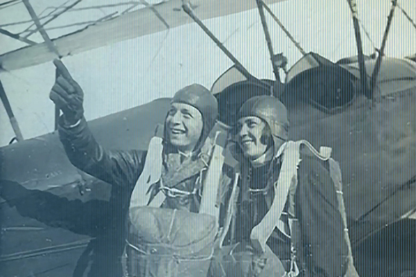 A historic photograph of a smiling man and woman wearing leather flight caps and parachute gear in front of a biplane, as the man points skyward.