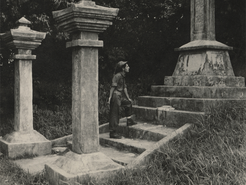 A black-and-white photograph showing a young US Navy Seabee standing on stone steps, looking upward at a Japanese stone memorial monument.