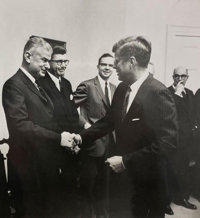 Black and white photo of 5 men in a room. Norman Obbard shaking hands with President John F. Kennedy.